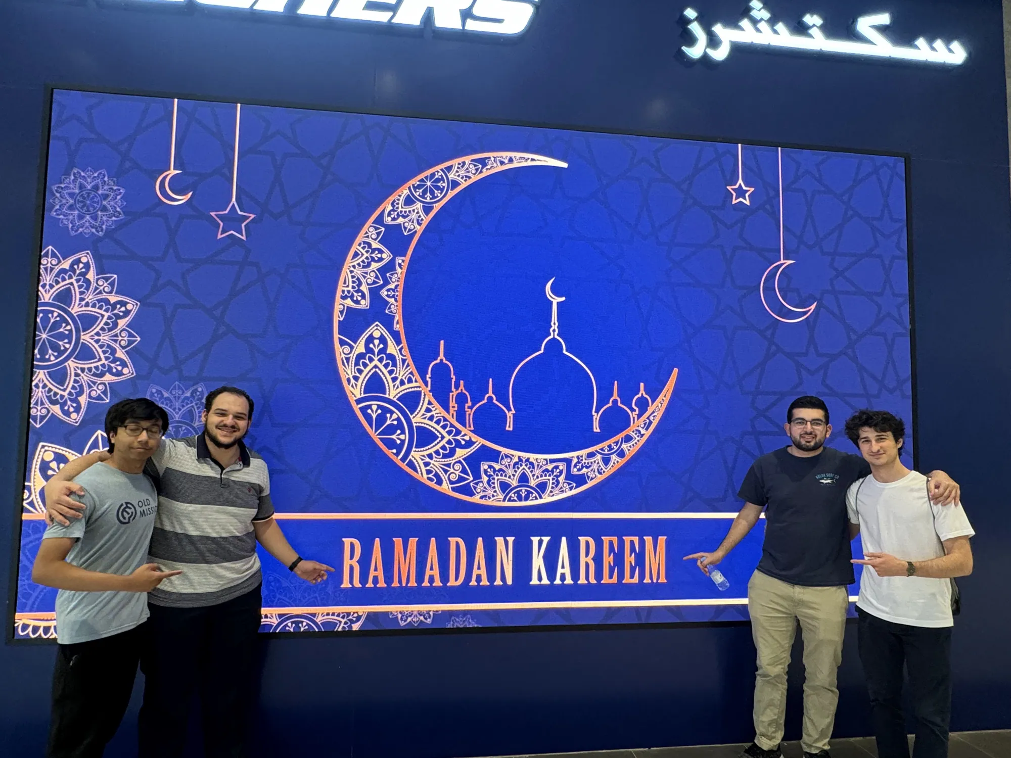 Students standing beside an illuminated Ramadan display in the evening.