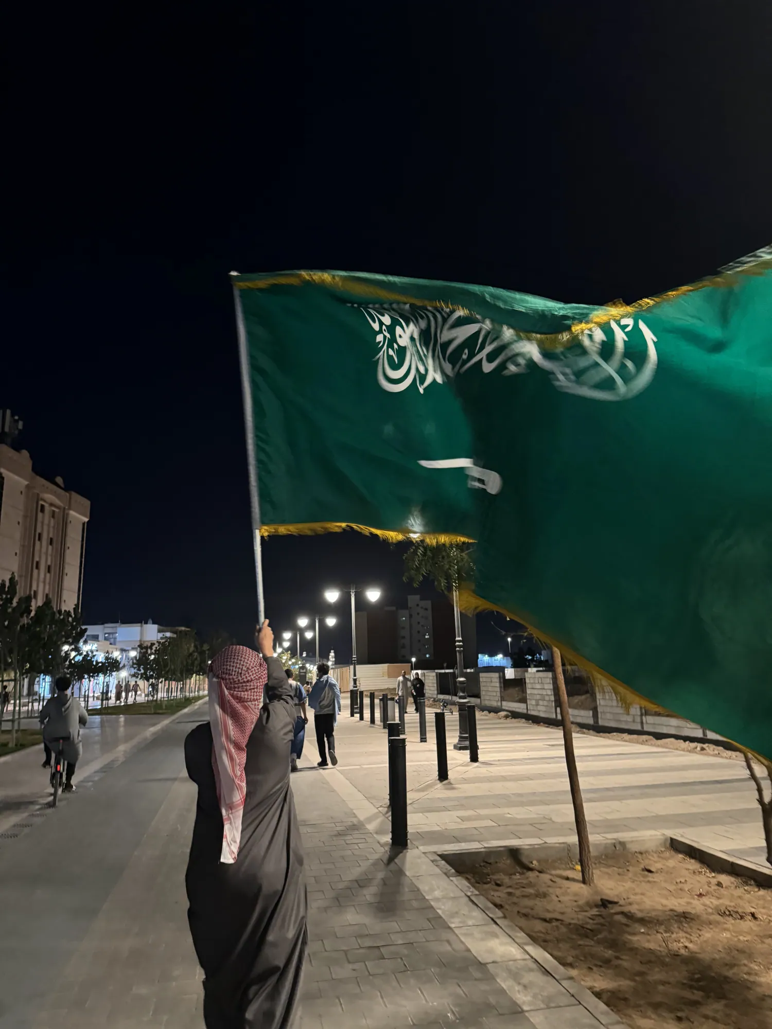 A student holding a Saudi flag during an evening walk in Madinah.